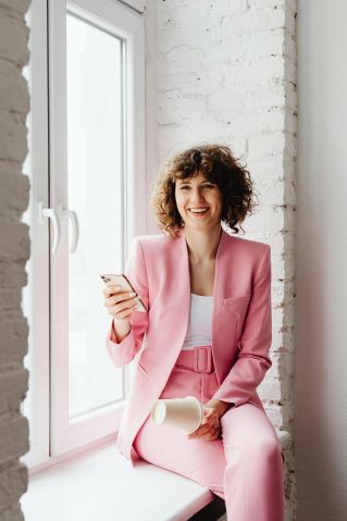 Brunette woman in a pink suit sitting by a window, smiling at her smartphone.